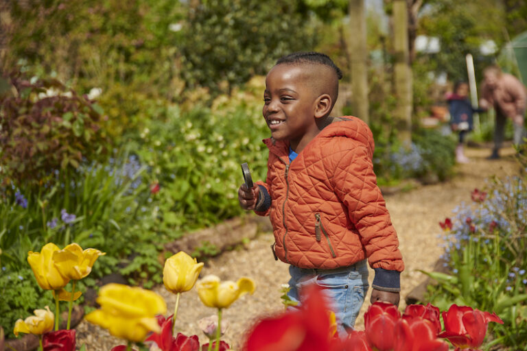 Finding great childcare in London can be a real challenge. The image shows a smiling child going on a nature hunt amongst flowers, holding a magnifying glass.