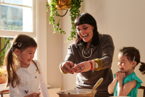 An experienced nanny cooks with two young girls she's taking care of.