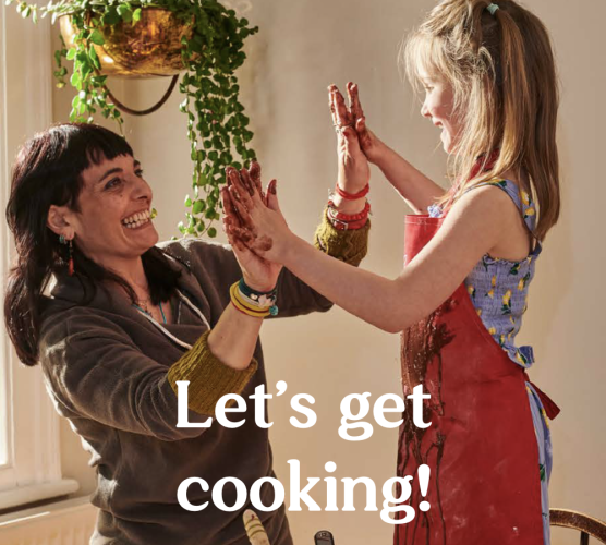 An experienced nanny is high-fiving the young girl she cares for while cooking. White text overlayed reads 'Let's get cooking!'